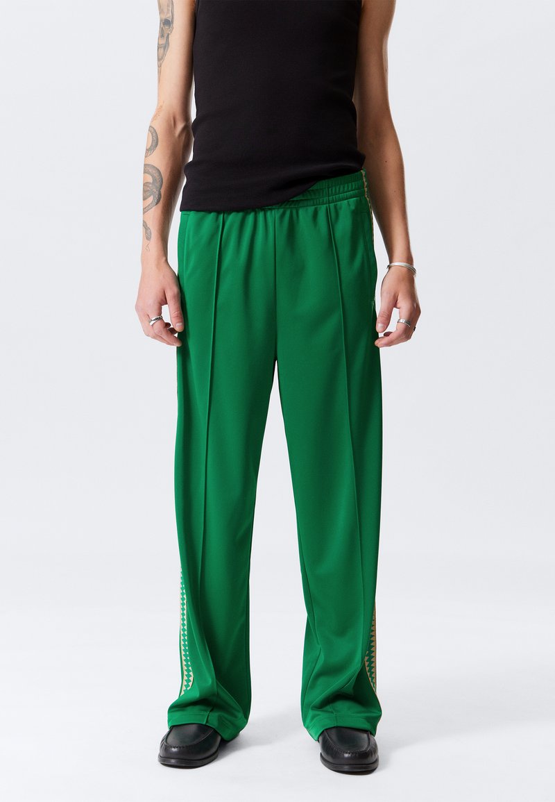 Groene sportbroek met bredere pijpen, een gladde textuur, een elastische tailleband en decoratief gele stiksels langs de zijnaden.