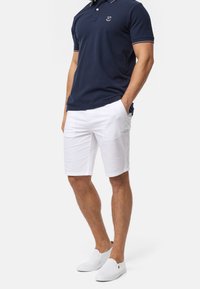 Polo shirt marine avec bord blanc, shorts blancs et chaussures slip-on blanches. Comprend un logo sur la poitrine. Tenue décontractée dans l'ensemble.