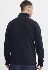 Svart fleece-genser med høy krage, lange ermer og rektangulær lappdetalj på baksiden. Myk tekstur og avslappet passform.