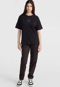 Sort bomulds T-shirt med et rundt blåt logo på brystet, parret med mørke cargo-bukser og hvide sneakers med sorte detaljer.