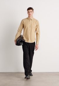 Beige button-up shirt with a spread collar, paired with black trousers. The model carries a black bag, standing on a neutral backdrop.
