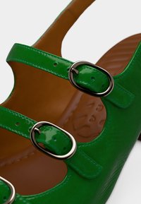 Chaussure en cuir verni vert avec trois brides réglables munies de boucles argentées, un bout arrondi et une semelle intérieure marron texturée.