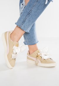 Baskets beiges avec finition brillante et larges lacets en satin blanc. Présentent des accents roses pâles et une semelle en caoutchouc texturé.