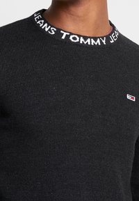 Suéter de punto negro con textura acanalada. El cuello presenta letras blancas "TOMMY JEANS." Pequeño logo en rojo y blanco en el pecho.