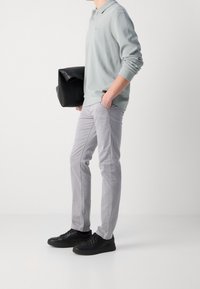 Jasnoszara koszulka polo z długim rękawem ze ściągaczami, zestawiona z jasnoszarymi spodniami o dopasowanym kroju oraz czarnymi sportowymi butami. W lewej ręce trzymana jest czarna torba.