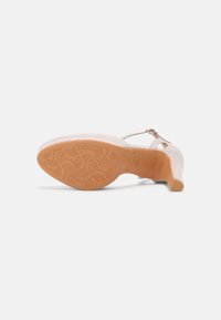 Sandale à talon haut en daim beige clair avec bride de cheville ajustable, semelle en caoutchouc texturée présentant un motif floral. Conception à bout fermé.