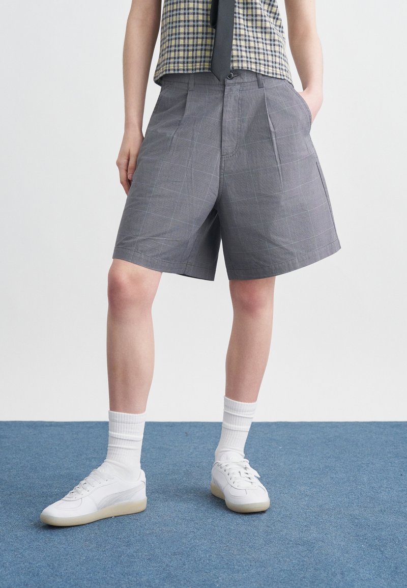 Άτομο που φοράει γκρι πλισέ σορτς, λευκά αθλητικά παπούτσια, λευκές κάλτσες και αμάνικο καρό τοπ με μαύρη γραβάτα, στέκεται σε μπλε χαλί.