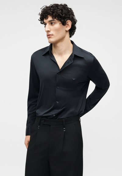 Νεαρός άνδρας με κατσαρά μαλλιά, που φοράει μαύρο πουκάμισο με κουμπιά και μαύρο παντελόνι, κοιτάζει προς τα πλάγια με το ένα χέρι πίσω από την πλάτη του.