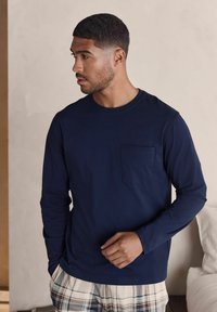 Navyblaues Langarmshirt mit rundem Halsausschnitt und Brusttasche. Aus weicher Stoffqualität; glatte Textur. Model trägt karierte Schlafanzughosen.