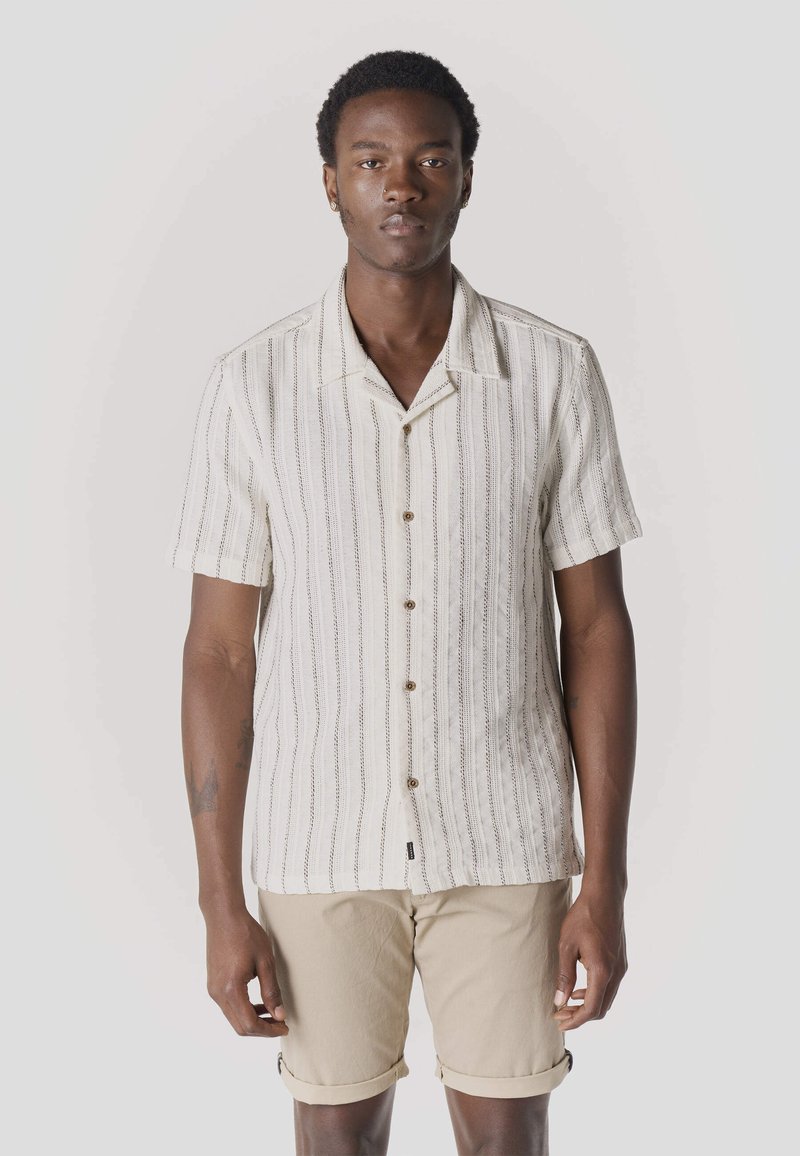 Mężczyzna ubrany w białą koszulę na krótki rękaw z pionowymi paskami oraz beżowe podwinięte szorty, stojący na jednolitym tle.