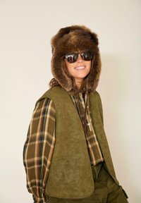 Gilet en daim vert porté sur une chemise à carreaux bruns et verts, associé à un chapeau doublé de fourrure et des lunettes de soleil noires, le tout sur fond neutre.