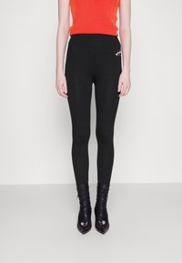 Leggings noirs au design ajusté, en matériau lisse, avec un petit détail de broderie rose. Associés à un haut rouge à côtes et des bottes noires.