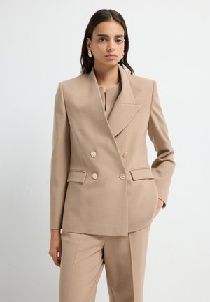 Beige double-breasted blazer with gold buttons and a structured fit. Textured fabric and notched lapels complement tailored pants.