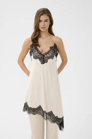 Kvinde iført en cremet slipkjole med sort blonderkant ved halsudskæringen og asymmetrisk kant, stående med hænderne bag ryggen på hvid baggrund.