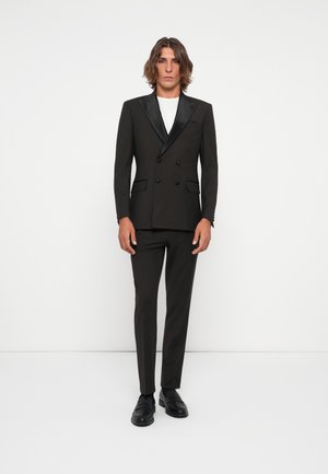 Svart dubbelknäppt kostym med satin-sjalkrage, slim-fit byxor och svarta loafers. Modellen har på sig en vit T-shirt under.