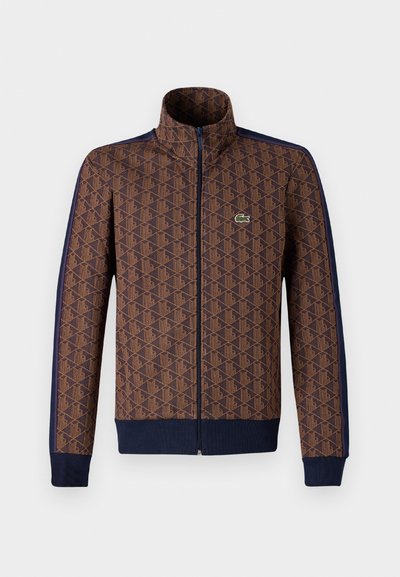 Veste zippée marron avec un motif géométrique, bandes latérales bleu marine, col montant et un petit logo vert sur la poitrine.