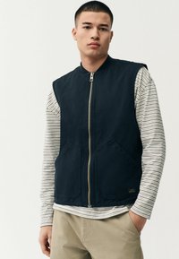 Marineblauwe mouwloze vest met een rits aan de voorkant, twee grote zakken en een lichte stof, gelaagd over een gestreept longsleeve shirt.