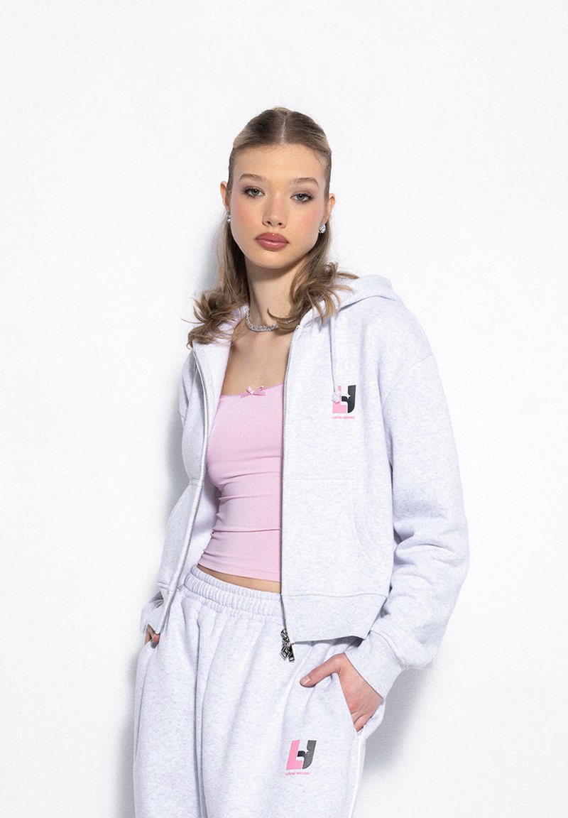 Jasnoszara bluza z kapturem na zamek, wykonana z miękkiego materiału, z różowo-czarnym logo. Nosi ją osoba w różowej bluzce typu crop top i pasujących spodniach.