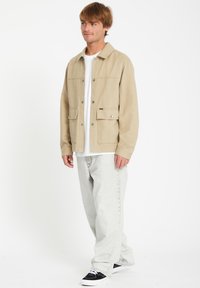 Chaqueta de lona beige con botones a presión, dos bolsillos frontales, llevada sobre una camisa blanca; combinada con jeans gris claro y zapatillas negras.