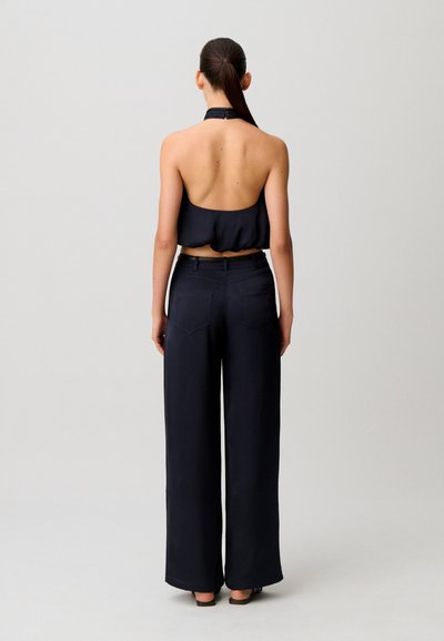 Donna con lunga coda di cavallo vista di spalle, che indossa un crop top nero senza maniche con scollo all'americana e pantaloni neri a gamba larga su uno sfondo semplice.
