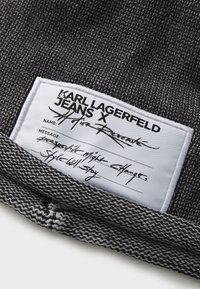 Grå vevd stoff med en hvit etikett som viser "KARL LAGERFELD JEANS X" og håndskrevet tekst, inkludert navn og en melding.