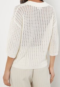 Lehký svetřík s otevřeným pletením v krémové barvě s žebrovaným límcem, tříčtvrtečními rukávy a texturovanými detaily. Nošený přes světle zbarvené kalhoty.