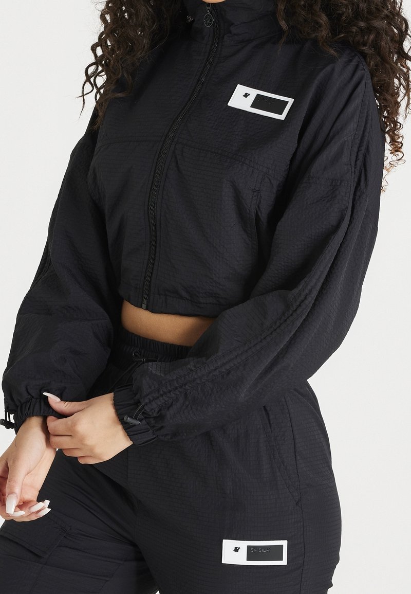 Femme portant une veste courte noire à fermeture éclair et un pantalon taille haute assorti avec des patchs de marque rectangulaires blancs.