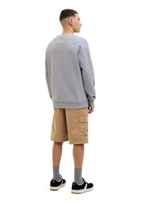 Grau Sweatshirt aus Baumwollmischung, kombiniert mit tanfarbenen Cargo-Shorts. Schwarze Sneaker mit weißen Akzenten. Modell von hinten gesehen.