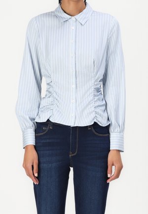 Jasnoniebieska koszula z długim rękawem na guziki, z białymi pionowymi paskami i marszczonymi bokami, w połączeniu z ciemnoniebieskimi dżinsami na stojącej osobie.