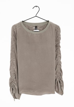 Blouse taupe à manches longues avec manches froncées nouées aux poignets, suspendue sur un cintre noir sur fond blanc.