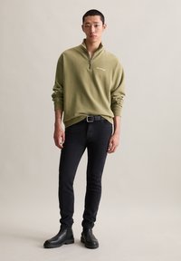 Man in olijfgroene kwart-rits sweatshirt, zwarte skinny jeans met riem, zwarte laarzen, staande tegen een effen lichte achtergrond.
