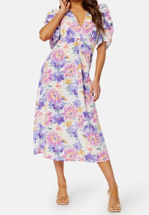 Femme portant une robe midi blanche à imprimé floral violet, rose et jaune avec des manches courtes bouffantes, associée à des talons à brides transparentes.