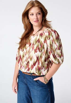 Femme aux longs cheveux roux portant un chemisier ample à motifs avec des manches bouffantes et un jean bleu, debout avec une main dans la poche.