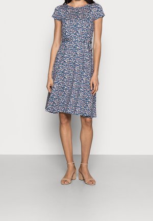 Woman wearing a knee-length blue floral dress with short sleeves and beige open-toe block-heeled sandals, standing on a gray floor.