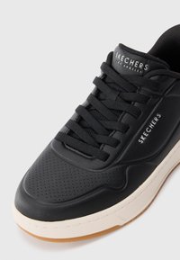 Black sneaker made of leather with a textured surface, featuring flat laces, a rubber sole, and a cream accent around the base. Brand logo visible.