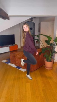 Nuori nainen viininpunaisessa neuleessa ja mustissa housuissa seisoo yhdellä jalalla olohuoneessa, jossa on puulattia, oranssi sohva, huonekasvi ja televisio.