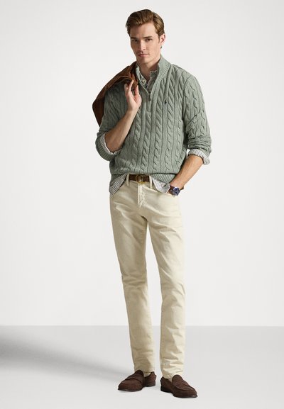 Light green cable-knit sweater over a checked shirt, beige chinos, and dark brown loafers, with a brown jacket draped over one shoulder.