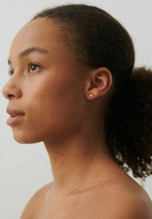 Fiatal nő göndör hajjal, visszafogva, balra néz, apró arany szív alakú bedugós fülbevalót visel, egyszínű világos háttér előtt.