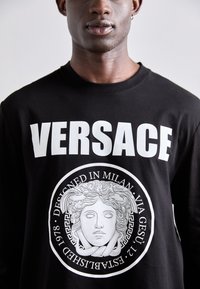 Melna kokvilnas krekls ar garām piedurknēm, kurā ir izteikta balta "VERSACE" teksts un centrāla grafika ar Medusas seju, ko ieskauj apļveida dizains.