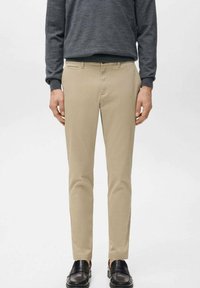 Mężczyzna ubrany w beżowe spodnie o dopasowanym kroju, ciemnoszarą koszulę z długim rękawem oraz czarne mokasyny, stojący na jednolitym jasnym tle.