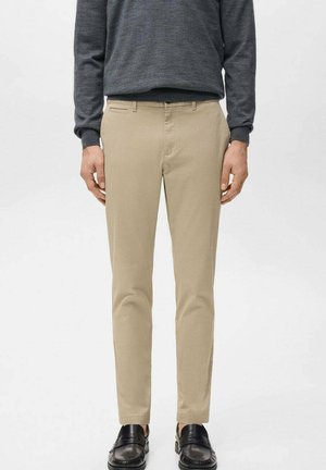 Man wearing beige slim-fit trousers, dark gray long-sleeve shirt, and black loafers, standing against a plain light background.