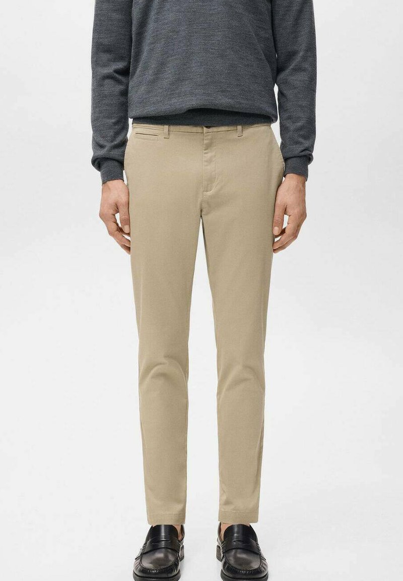 Mężczyzna ubrany w beżowe spodnie o dopasowanym kroju, ciemnoszarą koszulę z długim rękawem oraz czarne mokasyny, stojący na jednolitym jasnym tle.