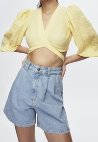 Haut jaune à manches bouffantes et tissu texturé, associé à un short en denim bleu clair taille haute avec fermeture à boutons et plis.