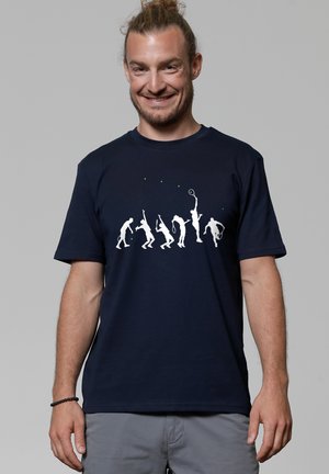 Homme souriant avec un chignon portant une chemise marine montrant six silhouettes blanches de joueurs de tennis en différentes poses d'action avec de petites balles de tennis vertes.
