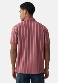 Bordová polo tričko s bielymi vertikálnymi pruhmi, krátkymi rukávmi a golierom. Textúrovaná látka; zadný pohľad zobrazuje spevnené švy.