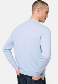 Mann mit kurzem Haar, der einen hellblauen Pullover und dunkle Jeans trägt, mit den Händen in den hinteren Taschen stehend, seitlich mit einem leichten Lächeln blickend.