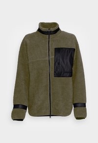 Giacca in pile verde oliva con una tasca sul petto nera con zip, colletto alto e polsini neri a contrasto. Morbido al tatto, vestibilità rilassata e zip completa.