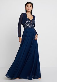 Abito da sera blu navy con lunghe maniche trasparenti, corpetto in pizzo e gonna fluida. Presenta dettagli con perline e un profondo scollo a V.