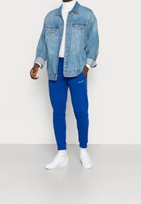 Jasnoniebieska kurtka dżinsowa z dwoma kieszeniami na piersi, pod spodem biały golf, niebieskie spodnie dresowe i białe trampki. Casualowy strój.