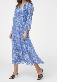 Pitkä sininen paisley-mekko, jossa pitkät hihat, V-kaula-aukko ja rypytetyt yksityiskohdat; kevyttä kangasta; yhdistetty mustiin avokärkisiin korkokenkiin.